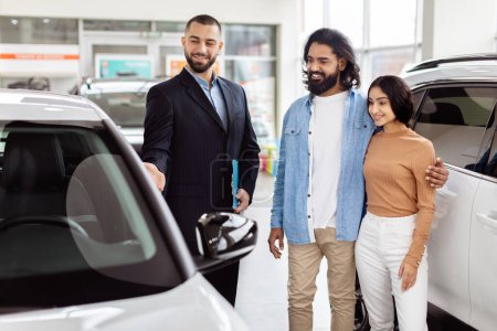 A young Indian couple is smiling and standing next to a new car at a dealership, interacting with a professional salesman who is offering the car keys
