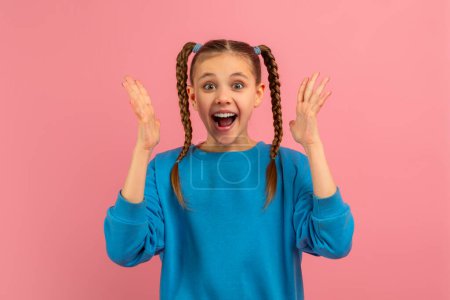 A young girl wearing a bright blue sweater stands against a vibrant pink backdrop, her hands raised in a gesture of surprise and elation