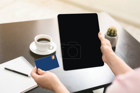 Recortado de mujer sentada en un escritorio, sosteniendo una tableta con una pantalla en blanco en una mano y una tarjeta de crédito en la otra, sugiriendo una sesión productiva o de planificación.