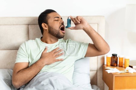 Afrikanischer junger Mann sitzt auf der Bettkante, hält sich mit einer Hand die Brust, während er Medikamente mit einem Asthma-Inhalator in seinen offenen Mund verabreicht