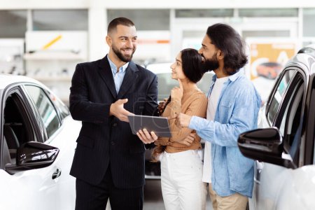 Photo for A cheerful salesman, holding a digital tablet, is engaging with Indian couple next to a new car inside a bright car dealership. They are in a conversation, possibly discussing the cars features - Royalty Free Image