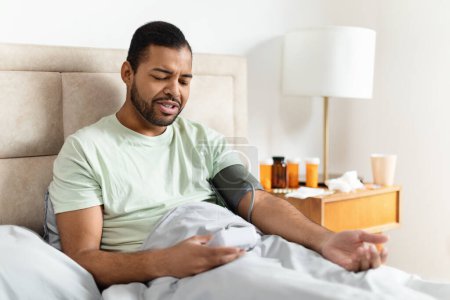 Ein junger Afroamerikaner sitzt im Bett, trägt ein lässiges T-Shirt und am Arm ein digitales Blutdruckmessgerät
