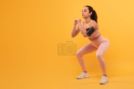 A young woman, dressed in activewear, is captured mid-motion as she performs a squat exercise, focused and maintains good form with her arms held in front of her, workout against yellow background