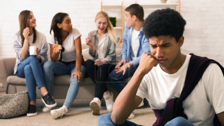 Photo for Black teenage boy appears visibly stressed and uncomfortable in the foreground while a group of his friends are engaged in lively conversation and laughter sitting on a couch in a well-lit room - Royalty Free Image