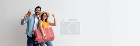 Family tours concept. Happy young couple with suitcase, passports and tickets posing over light wall and smiling at camera. Diverse tourists spouses ready for vacation trip, full length, copy space