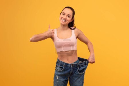 Eine fröhliche Frau mit breitem Lächeln steht vor leuchtend gelbem Hintergrund und hält ihre mittlerweile zu große Jeans hoch, um ihre beachtliche Gewichtsabnahme zu zeigen.