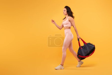 A young woman dressed in light-colored athletic wear confidently strides forward, carrying a black and red gym bag, copy space