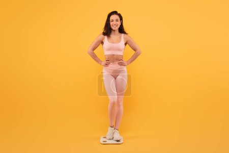 A smiling young woman stands on scale with her hands on her hips, wearing light pink activewear and white sneakers. She exudes confidence and positivity against a vibrant yellow backdrop