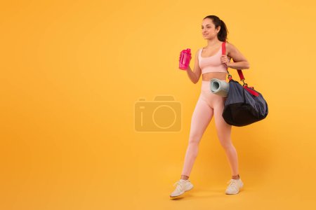 Téléchargez les photos : Une jeune femme est habillée en tenue d'entraînement, y compris un soutien-gorge de sport et des leggings, portant un sac de sport noir sur son épaule. Elle tient une bouteille d'eau rose et semble être en mouvement, copier l'espace - en image libre de droit