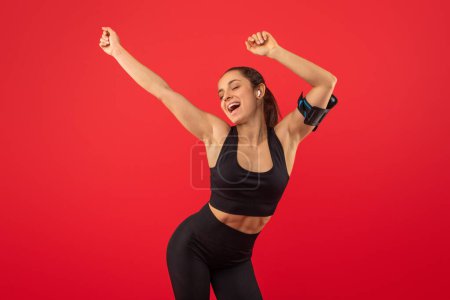 A young woman in sportswear, with her arms raised in victory, exudes joy and accomplishment against a vibrant red backdrop, reached a personal fitness milestone or achieved success in workout routine