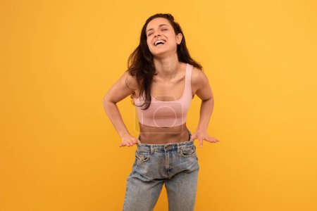 Foto de Una joven alegre vestida con una camiseta sin mangas de color rosa pastel y vaqueros azules se levanta contra un fondo amarillo vivo, con las manos en las caderas mientras inclina la cabeza hacia atrás en una risa espontánea - Imagen libre de derechos