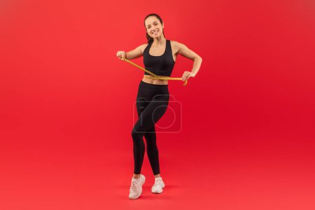 Eine fröhliche junge Frau steht vor leuchtend rotem Hintergrund und hält ein Maßband um ihre schlanke Taille, um ihren Fitness-Fortschritt zu demonstrieren.