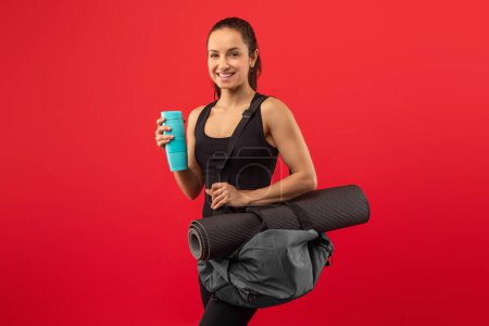 A woman is standing while holding a yoga mat in one hand and a cup in the other hand. She appears to be ready for a yoga session or workout, balancing the items with ease.