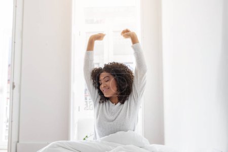 Hispanic woman is waking up in bed, stretching with her arms raised up in the air. She is lying down on a white bedsheet with pillows in the background.