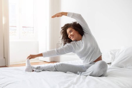 Hispanic woman is stretching her body while lying on a bed with white sheets. She is extending her arms and legs to loosen up her muscles after waking up.