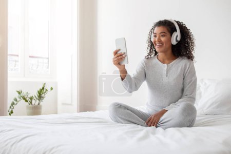 Una joven hispana alegre con el pelo rizado está sentada con las piernas cruzadas en una cama blanca, sosteniendo su teléfono inteligente para una videollamada. Ella está usando ropa casual acogedora y auriculares blancos