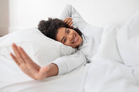 A cheerful Hispanic woman is lying in bed, with her head resting on a pillow and a comfortable smile on her face. She is casually dressed in cozy, light sleepwear, reaching out towards the camera