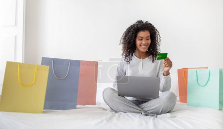 Una alegre joven hispana está sentada en la cama con las piernas cruzadas, rodeada de coloridas bolsas de compras, sosteniendo una tarjeta de crédito en la mano mientras mira la pantalla de su computadora portátil