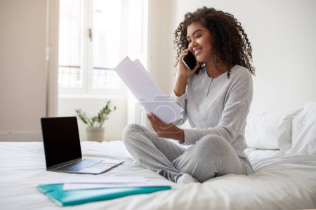 Hispanic young woman, comfortably dressed in casual athleisure wear, sits cross-legged on a white bedspread. Engaged in a phone conversation, she holds a document in one hand with a laptop open