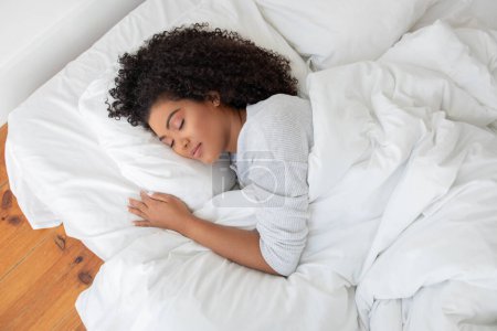 La mujer hispana está acostada en una cama con un edredón blanco cubriéndola, luciendo relajada y cómoda en un ambiente de dormitorio, vista arriba