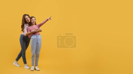 Mother and daughter dressed in casual wear, stand closely together against a vibrant yellow backdrop, pointing at copy space while they both smile brightly, sharing a lighthearted moment.