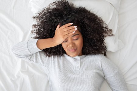 Una joven hispana está acostada boca arriba en una cama blanca con los ojos cerrados y una expresión dolorosa, presionando su mano contra su frente, sufriendo de un dolor de cabeza o sintiéndose mal.