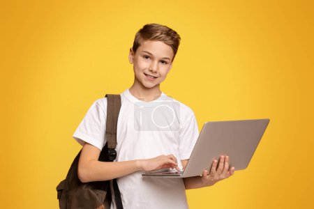 Téléchargez les photos : Jeune garçon prêt pour l'école. Adolescent avec sac à dos à l'aide d'un ordinateur portable, fond panoramique orange avec espace libre - en image libre de droit