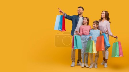Une famille joyeuse, avec deux parents et deux enfants, est debout ensemble tenant des sacs à provisions colorés. Le père fait remarquer quelque chose au loin, copier l'espace