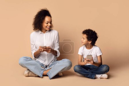 Mujer afroamericana y un niño pequeño están sentados con las piernas cruzadas uno frente al otro contra un telón de fondo beige. Madre sonríe y mira su teléfono inteligente, mientras su hijo sostiene otro teléfono inteligente
