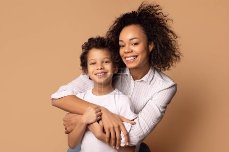 A joyful African American mother is tightly hugging her curly-haired young son, who radiates happiness. They are both smiling genuinely, as they share a special moment of connection and love