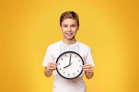 A young boy holds a clock in front of his face, showcasing the time with a serious expression.