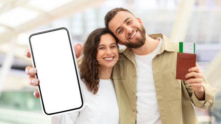 Foto de A cheerful man and woman are shown close together holding up a smartphone with a blank screen and a passport. They seem to be in a well-lit airport, possibly ready for travel - Imagen libre de derechos