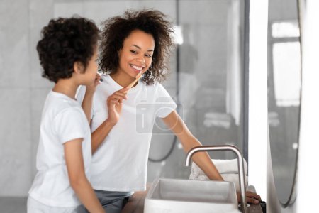 Black woman is seen standing next to a young boy, both brushing their teeth. Mother holds a toothbrush and looks at her reflection in the mirror, son mimics her actions