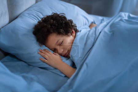 Téléchargez les photos : African American jeune garçon dort paisiblement dans un lit avec des draps bleus. Il a l'air confortable et détendu, les yeux fermés et le corps dans une position reposante. - en image libre de droit