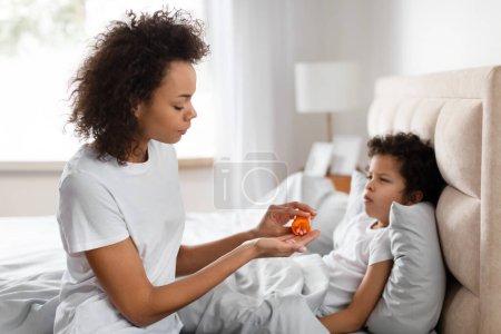 Una madre afroamericana preocupada se sienta al lado de su pequeño hijo postrado en cama, administrando cuidadosamente medicamentos orales. La luz natural de una ventana cercana ilumina el dormitorio sereno