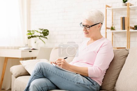 Die Seniorin sitzt auf einer Couch und schreibt konzentriert auf einen Zettel, der auf ihrem Schoß liegt. Ihr Fokus ist intensiv und unterstreicht ihr Engagement für die bevorstehende Aufgabe..
