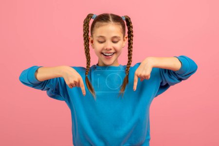 Téléchargez les photos : Une jeune fille joyeuse avec deux longues nattes tressées porte un pull bleu vif. Elle se tient contre une toile de fond rose solide, pointant vers le bas avec ses deux index - en image libre de droit