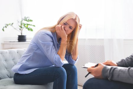 Una mujer se sienta en el borde de un sofá, su mano cubriéndose la cara, posiblemente secándose las lágrimas, expresando angustia o frustración durante una sesión de asesoramiento