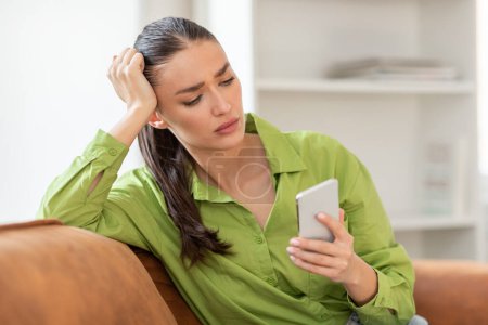 A woman sitting on a couch indoors, engrossed in her cell phone screen. She appears focused as she scrolls and interacts with the device, seemingly unaware of her surroundings.