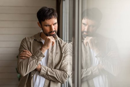 Pensive millennial bearded man standing next to window at home, touching his chin, deep in his thoughts, experiencing difficulties, copy space