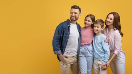 Foto de Una familia feliz, con dos padres y dos hijos, se abrazan contra un vibrante telón de fondo amarillo. Aparecen alegres y contentos, mirando el espacio de copia - Imagen libre de derechos