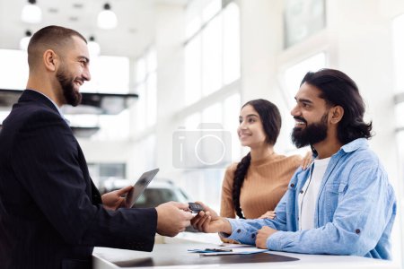 A joyful Indian couple stands at a dealership counter, receiving a new set of car keys from a professional salesman. The showroom is well-lit