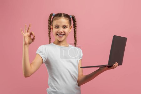 Photo for A young girl is standing against a pink background, holding a laptop computer in her hands. She appears to be engrossed in the screen, possibly playing a game or watching a video - Royalty Free Image