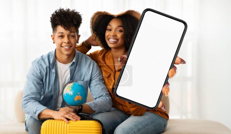 Téléchargez les photos : Un jeune couple noir souriant s'assoit étroitement ensemble, tenant un smartphone à écran blanc adapté aux maquettes, suggérant qu'ils planifient un voyage ou des vacances. - en image libre de droit