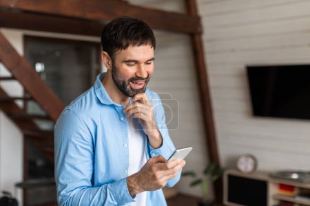Un joven alegre con barba se ve sosteniendo y mirando su teléfono inteligente con una sonrisa, lo que sugiere que ha recibido buenas noticias