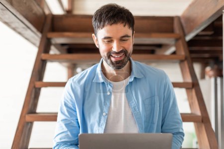 A cheerful young man with a beard is focused on his laptop screen while working indoors, seated at a wooden stairs in a well-lit, airy space that suggests a home or a casual office environment.