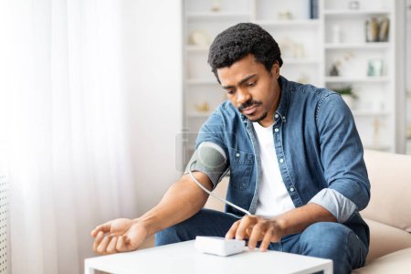 Ein schwarzer Mann sitzt auf einer Couch und konzentriert sich darauf, seinen Blutdruck mit einem digitalen Blutdruckmessgerät zu messen. Sein Arm ist in die Manschette gewickelt und mit einem kleinen, tragbaren Gerät verbunden.