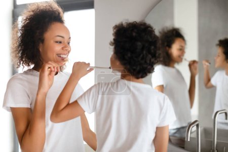 Foto de Mujer joven afroamericana con una sonrisa brillante se cepilla los dientes frente a un espejo de baño, vestida con una camiseta blanca casual y participa activamente en su práctica diaria de higiene bucal. - Imagen libre de derechos