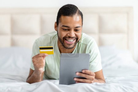 Téléchargez les photos : Afro-Américain est allongé sur un lit tout en tenant une carte de crédit dans une main et une tablette dans l'autre, éventuellement se livrer à des achats en ligne ou la gestion des finances. - en image libre de droit