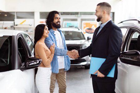 Ein junges indisches Paar schließt gerade einen Deal mit einem Autoverkäufer in einem Autohaus ab. Sie wirken zufrieden und schütteln dem Agenten, der im Businessanzug auftritt, die Hand.